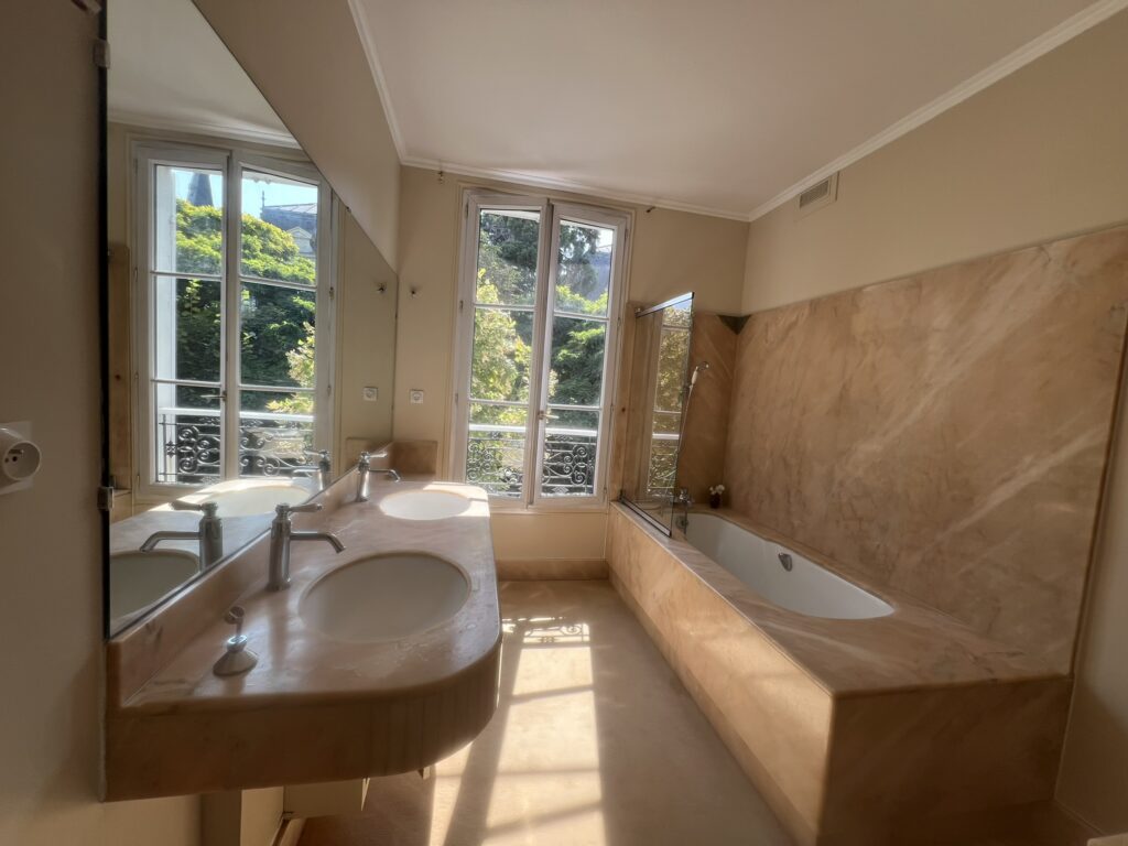Salle de bain rénovée d’une maison de style château à Enghien-les-Bains – murs peints en ton beige doux, revêtements effet marbre, double vasque et baignoire lumineuse